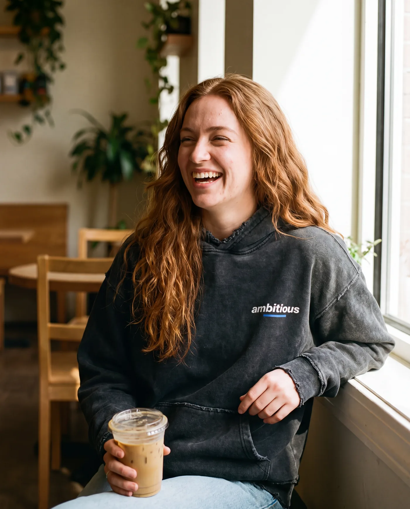Woman laughing in a café wearing the hoodie, ambitious chest logo